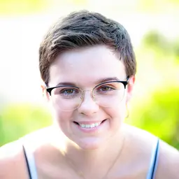 Nahaufnahme einer freundlich lächelnden jungen Frau mit kurzen Haaren und modischer Brille vor hellem, verschwommenem Hintergrund lens: 56mm F1.4 DC DN | Contemporary 018