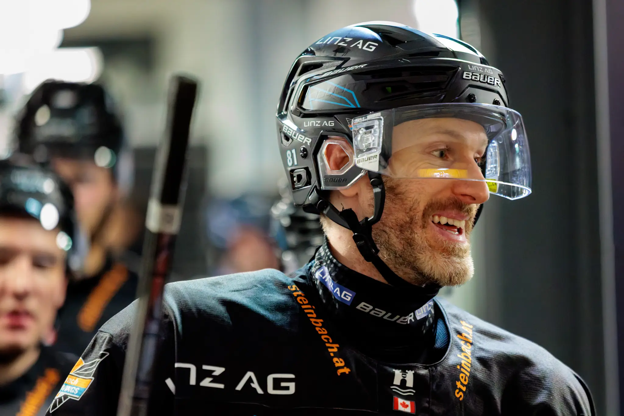 Nahaufnahme des lächelnden Eishockeyspielers Sean Collins von den Steinbach Black Wings Linz in seiner schwarzen Ausrüstung mit Helm